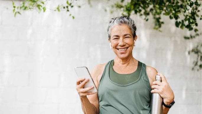 A senior woman wearing activewear is holding a phone and a water bottle as she exercises outdoors