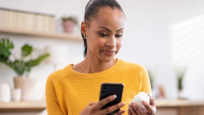  A young woman wearing a yellow top is holding a bottle of pills in one hand and her phone in the other hand