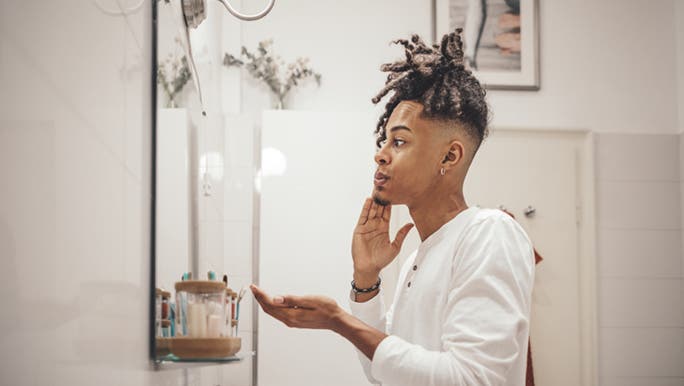 In a neutral bathroom, a man with dreadlocks puts cream on his face, wondering if sensitive skin is a skin type or condition.
