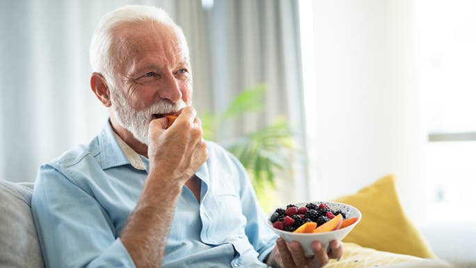 A senior man wearing a blue shirt is sitting on a sofa eating a bowl of fruit that’s good for gut health, including berries