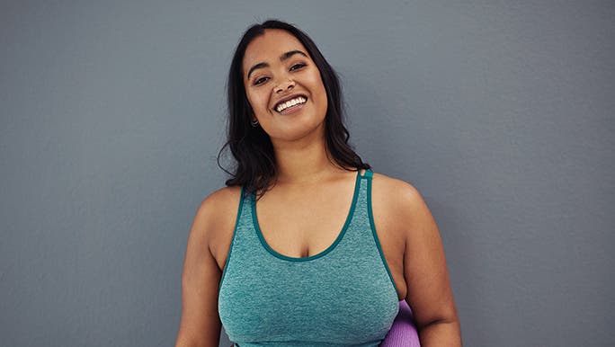 A lady with a yoga mat smiles at the camera. She is wearing activewear and standing in front of a smooth grey wall. 
