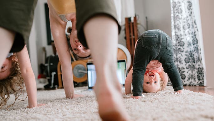 From a low and crooked angle, a baby us bending over and looking through her legs. Her mum is doing the same thing. It looks like the family is doing a fitness challenge that includes yoga. 