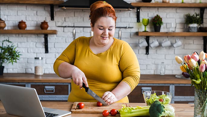 A woman is at a kitchen bench cutting vegetables with her laptop open, reading about foods that reduce cortisol.