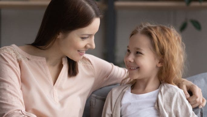 A young mother is sitting beside her young daughter and smiling as she holds her arm around the child’s shoulder