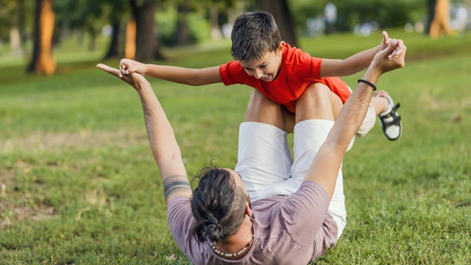 A father and his son are outdoors on the grass. His son is in a plane position, with his stomach safely resting on his fathers knees. His father is lying on his back to support his son, both happy to be outside. Laughter and nature are ways to feel good.
