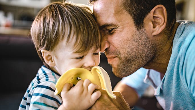 A man is sitting close to a young boy who is eating a banana, a snack food considered good for gut health