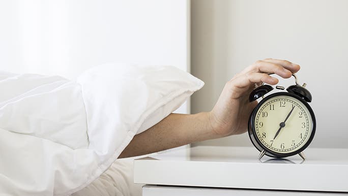 From under a white quilt, a hand reaches out to turn off a vintage alarm clock after reading how to catch up on sleep.
