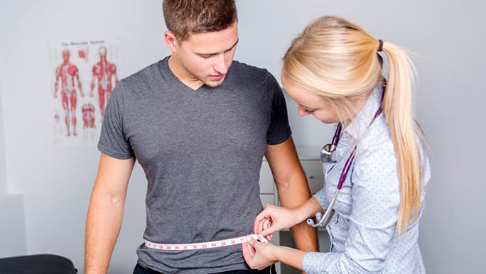 A young blonde female medical professional is measuring a young man’s waist circumference
