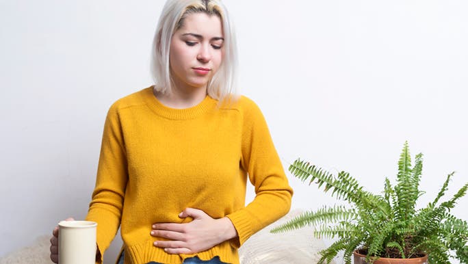 A young blonde woman wearing a yellow jumper is sitting down holding a cup of coffee and holding her stomach