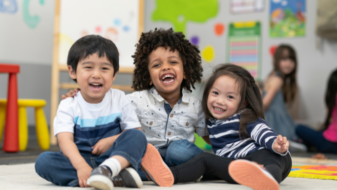 Three toddlers are sitting on the floor at daycare and smiling as they hug each other
