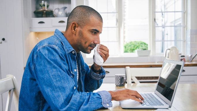 A young black man wearing a denim shirt is wiping his nose while working on a laptop