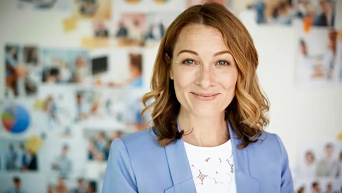 Woman in a white top and blue jacket smiling infront of a wall of pictures