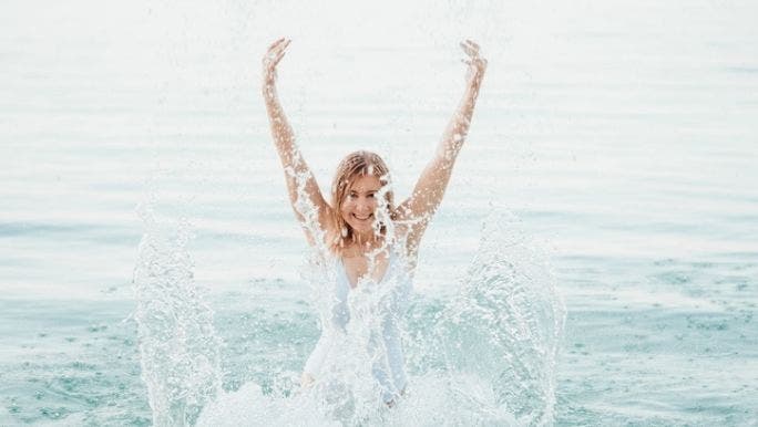 A woman is wearing a white bathing suit and smiling as she splashes in a body of water with her arms in the air