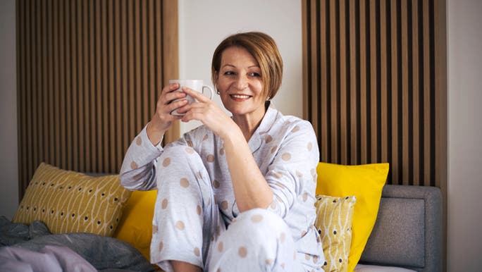 A woman is smiling and sitting in bed wearing pyjamas and holding a cup of coffee