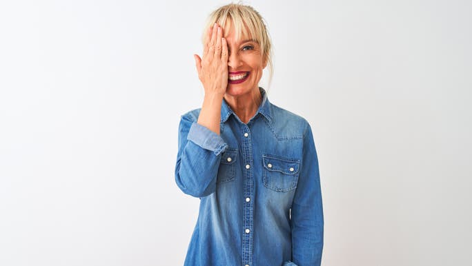 Middle age woman wearing casual denim shirt standing covering one eye with hand, confident smile on face and surprise emotion