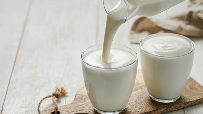 On a table, two glasses of yogurt stand on a wooden chopping board. One is still being poured.
