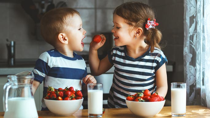 Two toddlers a boy and a girl in blue and white stripe tops eating strawberries in the kitchen