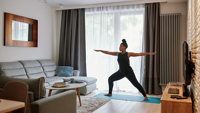A woman is doing yoga in her lounge room, she is doing warrior one position. 