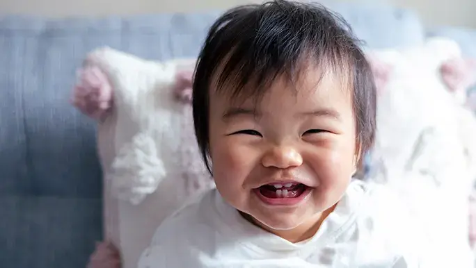 Baby is sitting on the couch smiling to camera showing off their baby teeth.