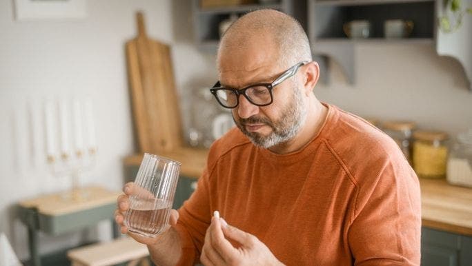 A middle-aged man wearing black glasses is holding a glass of water and looking at a white pill
