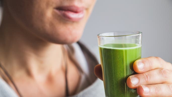 A  close up of a woman in a grey jumper holding a glass of the water mixed with the best greens powder in Australia.