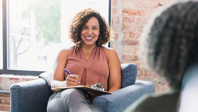 A dietitian is sitting on an armchair doing what a dietitian does, consulting with a patient. She has a big smile and curly brown hair. 
