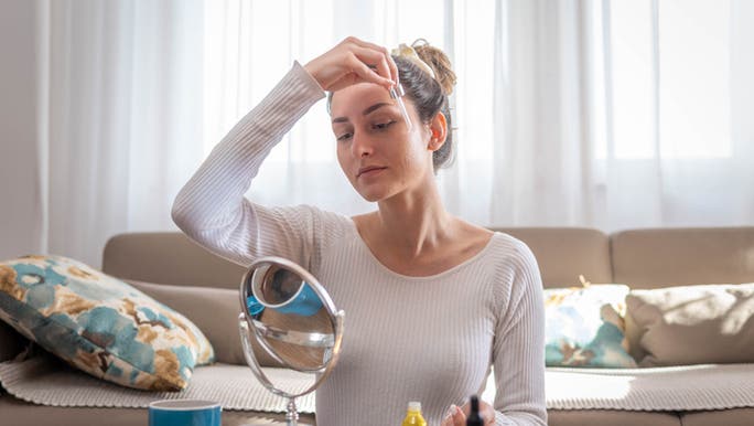 A young woman is looking into a mirror and applying a skin serum to her face