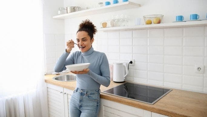A young woman is standing in a kitchen eating from a white bowl