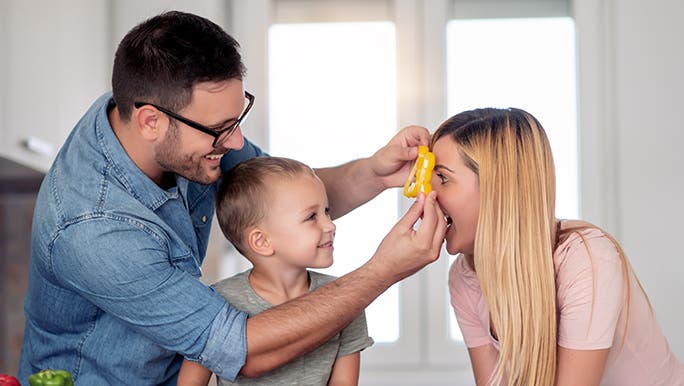A child is watching his father hold a vegetable piece up to his mother’s eye as she plays along with the game as a way of making vegetables fun.  