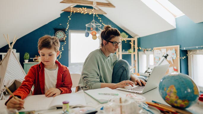 Mum working from home on a laptop next to her son doing his schoolwork
