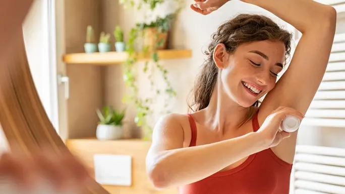 Woman with curly brown hair in a red singlet applying natural deodorant in the mirror