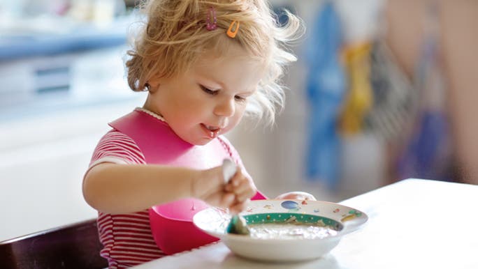 A toddler eats a bowl of oats at a kitchen bench. She has a pink rubber bib on. 