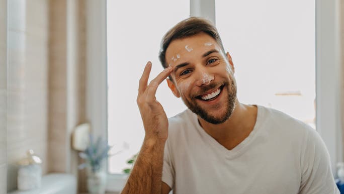 A young Caucasian man wearing a white T-shirt is smiling and applying a skincare product to his face