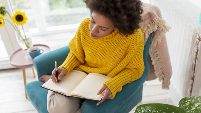 A lady in a bright yellow jumper is sitting on a blue armchair and updating her daily planner.