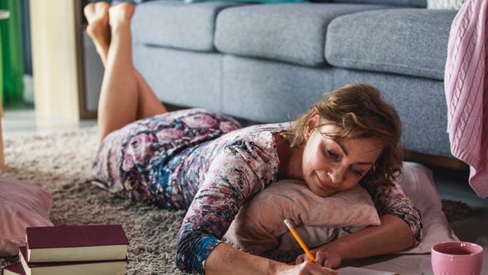 A lady lays on the floor next to a couch, she is writing her to-do list and it’s full of productivity hacks. 