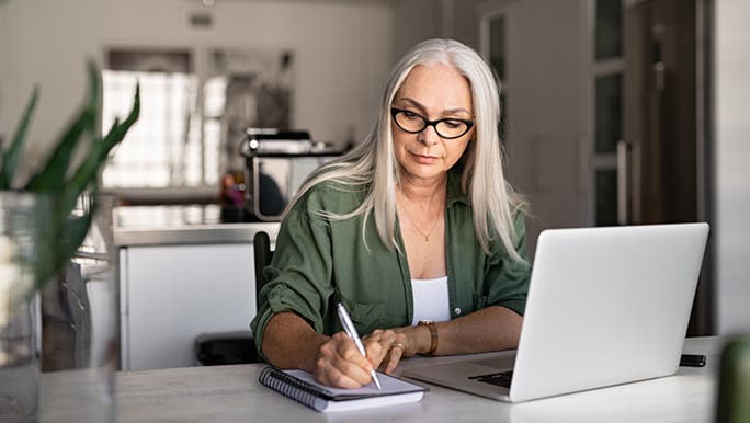 A woman with long grey hair is sitting at a desk in front of a laptop. She is writing in her daily planner/diary. 
