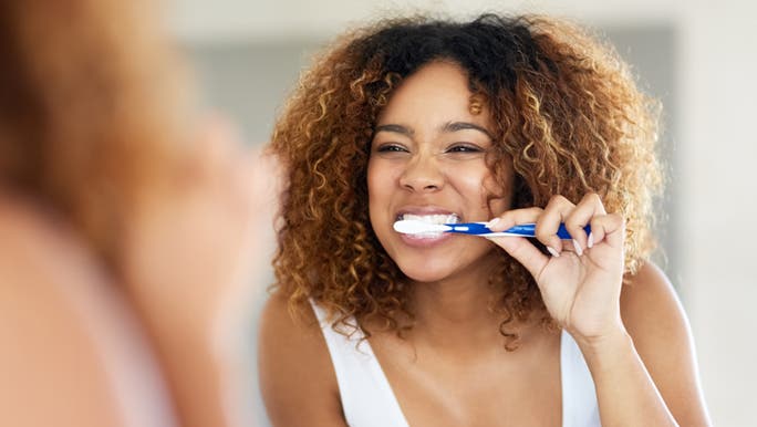 A woman smiles into the mirror while cleaning her teeth as she knows how best to brush them. 