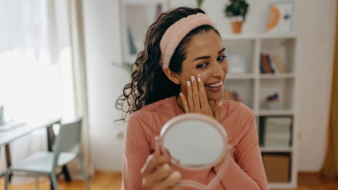 A young woman is smiling into a handheld mirror as she applies face cream