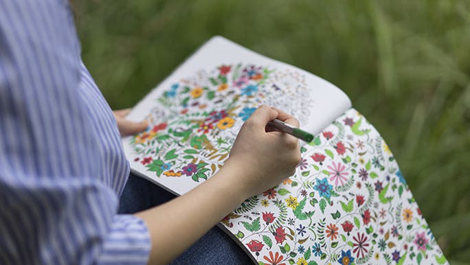 A lady sits outside in the grass, balancing a mindfulness colouring book on her knees.