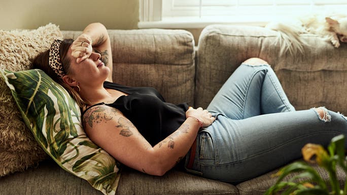 A woman wearing jeans and a black top is laying on a couch and touching her stomach