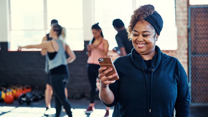 A woman is smiling whilst she looks down at her phone, she just finished her workout and is excited to know this is good for her well-being.