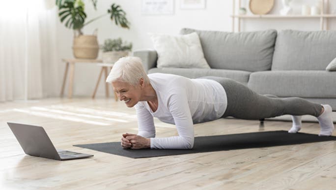 A woman with short grey hair does a plank on her lounge room floor while watching a video on her laptop about functional training exercises.