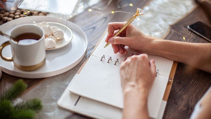 A woman sits at a desk with a cup of tea, writing in her journal after pondering the meaning of a new years resolution.