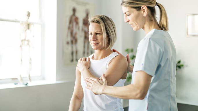 A woman is stretching her shoulder with the help of a physiotherapist. They may be discussing why stretching is important. 