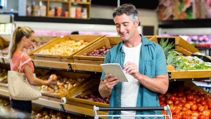 A middle-aged man is shopping in a grocery store and looking at a shopping list on a notepad