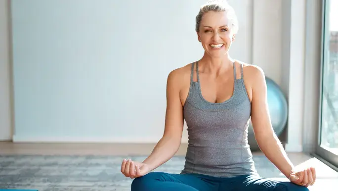 A woman is dressed in yoga gear and sitting down on the floor upon a yoga mat engaging in a yoga post. She is smiling towards the camera.