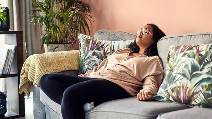 A woman with a dark complexion and wearing glasses is resting on a grey sofa