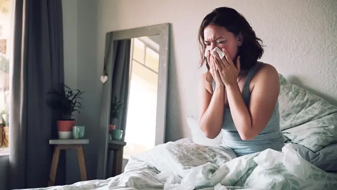 Woman suffering with the common cold and sneezing into a tissue while sitting on her bed at home