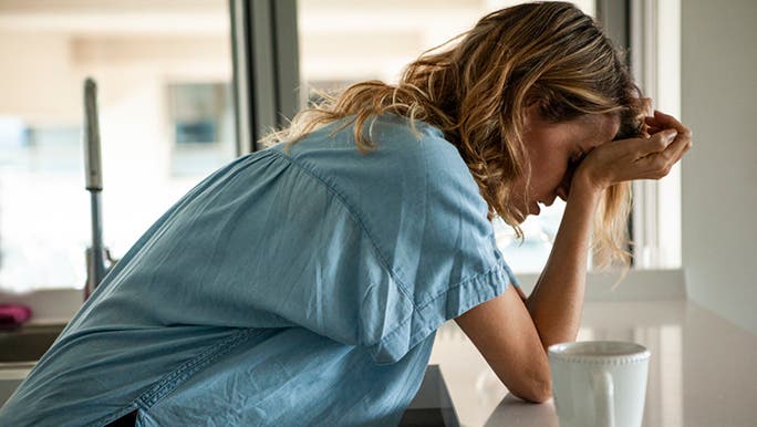 A woman wearing a blue shirt is leaning on a kitchen counter holding her head with a coffee mug and pain medication beside her