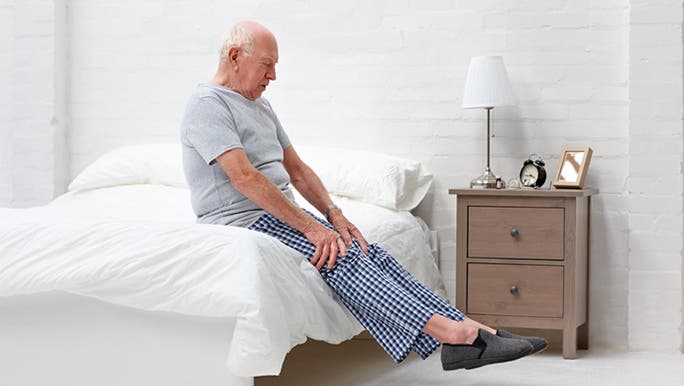 An elderly man is sitting on the edge of a bed in a bright room and stretching his legs
